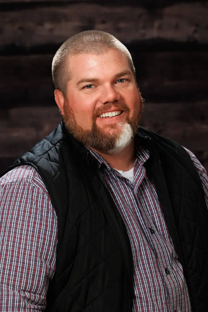 professional headshot of the founder of 1805 Consulting, smiling and wearing a plaid shirt and dark vest in front of a wood background